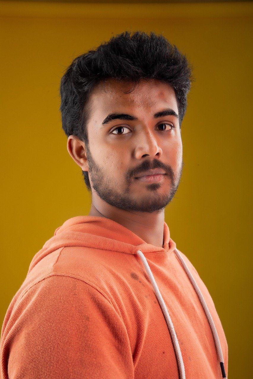 A young Indian man wearing an orange hoodie, looking confidently at the camera against a solid yellow background.