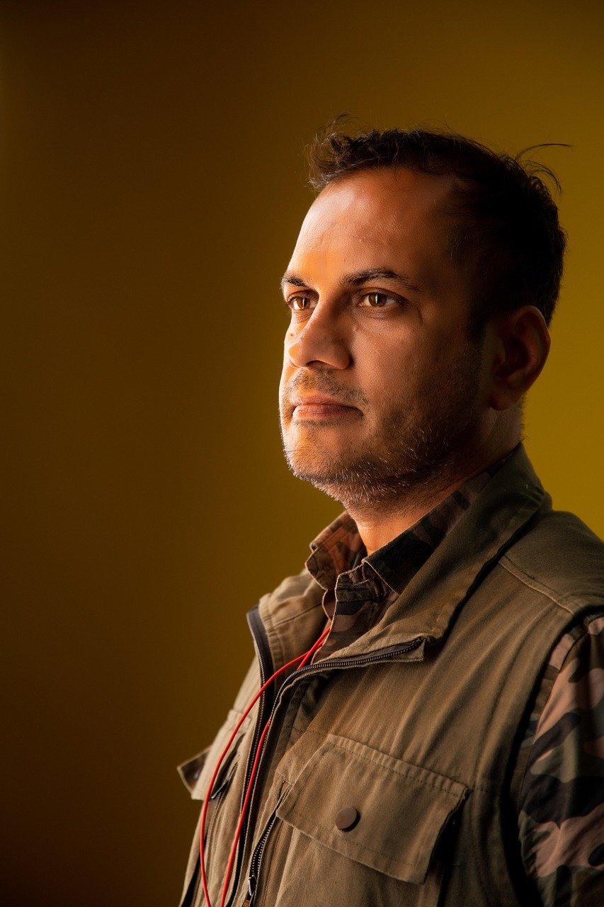 A side-profile portrait of an Indian man wearing an olive green jacket, looking thoughtfully into the distance against a warm yellow background.