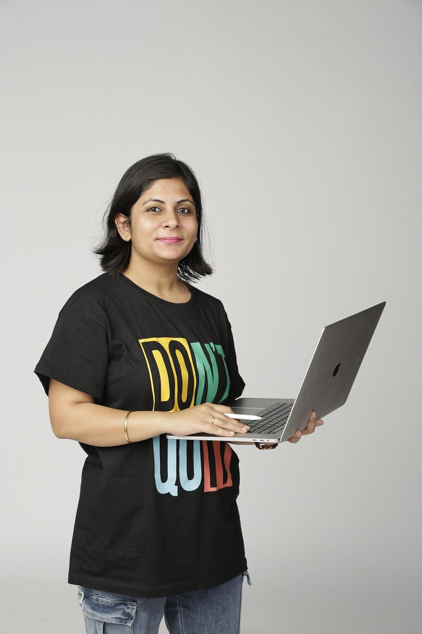 An Indian woman confidently holding a laptop, wearing a black "DON'T QUIT" T-shirt and jeans, standing against a plain light background.